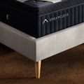 High-angle profile shot showing the edge of the DreamCloud Luxe mattress on an upholstered bed frame with natural wood tapered legs.
