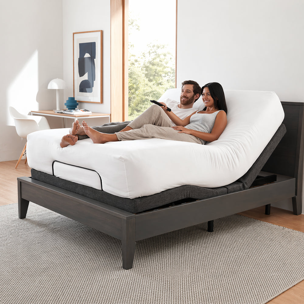 Happy couple relaxing on a SleepTone S1000 adjustable bed base inside a dark wood frame, using the head elevation feature for ergonomic TV viewing.

