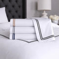 Stacked SleepTone Hotel Luxury sheets in white with gold, silver, and navy embroidered borders.
