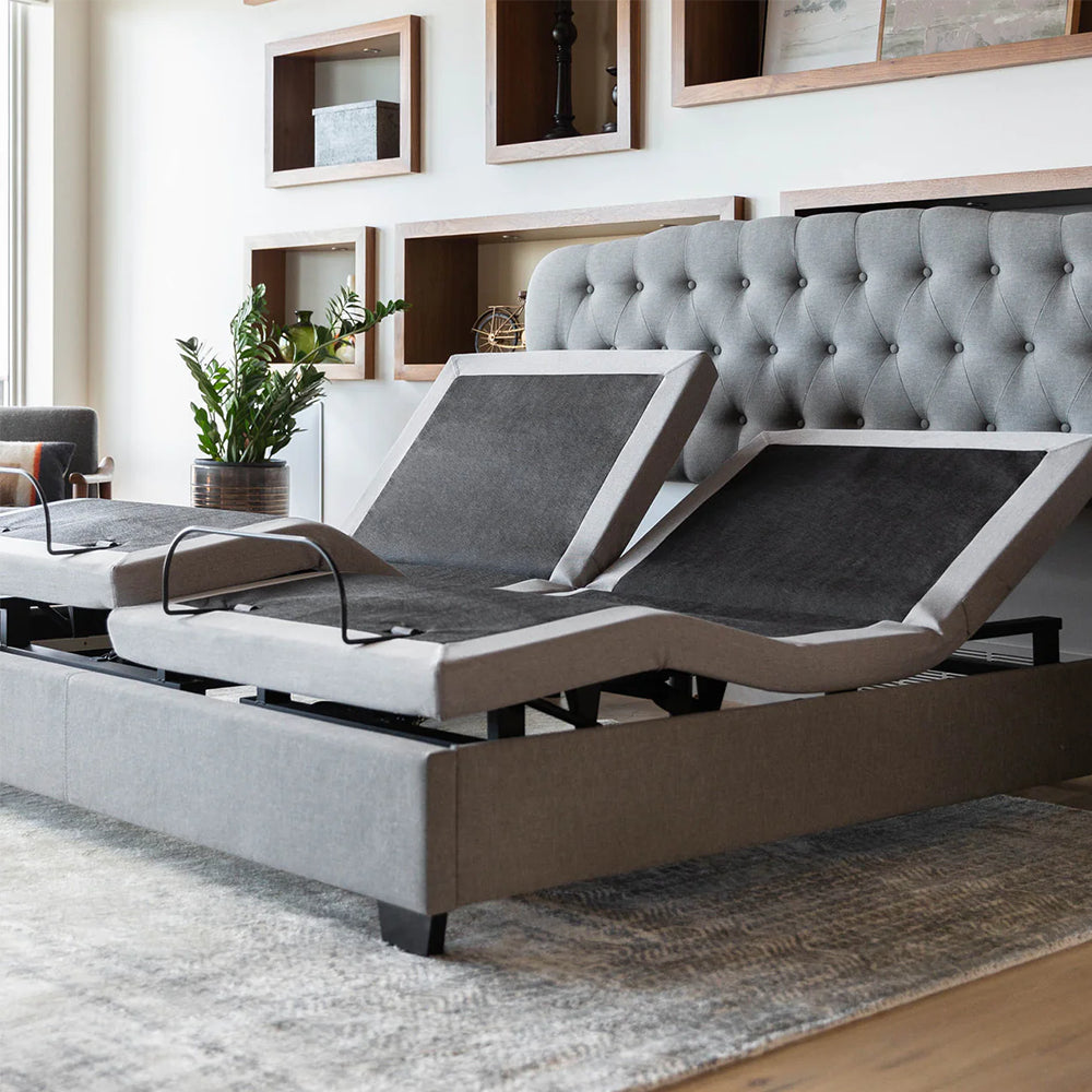 Split king Malouf M555 adjustable bed base in a lifestyle bedroom with a grey tufted headboard and articulated head sections.
