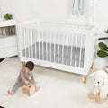 Baby playing near a white crib with a grey moisture-wicking Dri-Tec performance crib sheet.
