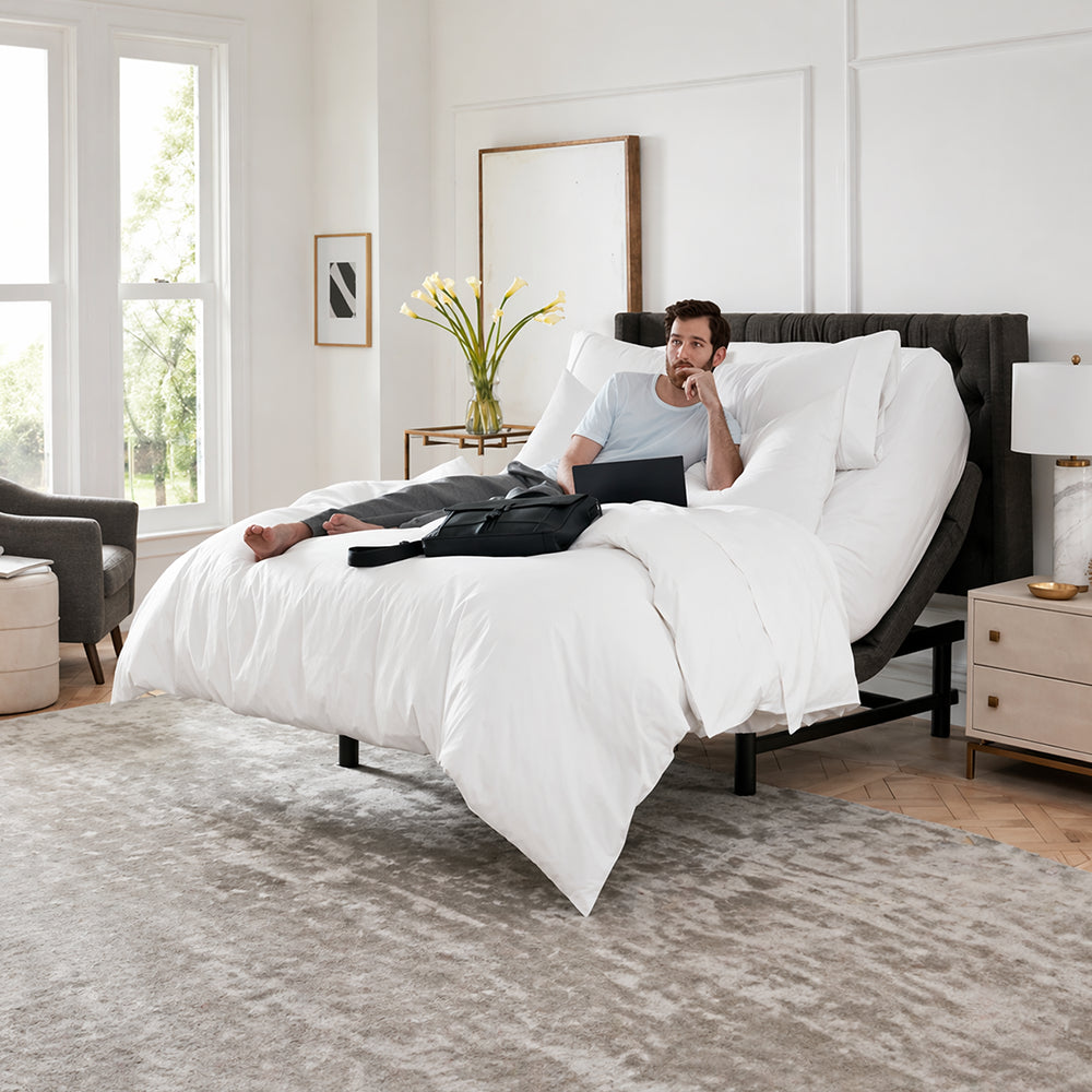 Man using a laptop while resting on a SleepTone S5000 adjustable motorized base, demonstrating ergonomic head elevation for home office comfort.
