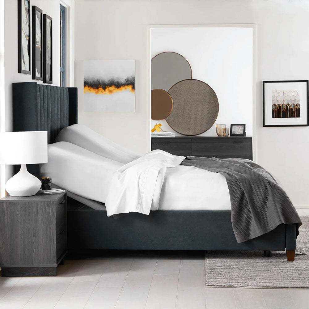 Side view of the Malouf S755 power base with head section elevated, shown with a mattress and white bedding in a contemporary room.

