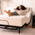 Lifestyle image of a woman relaxing on an MLILY MS 200 adjustable bed base with a mattress, showing head and foot elevation for lounging.

