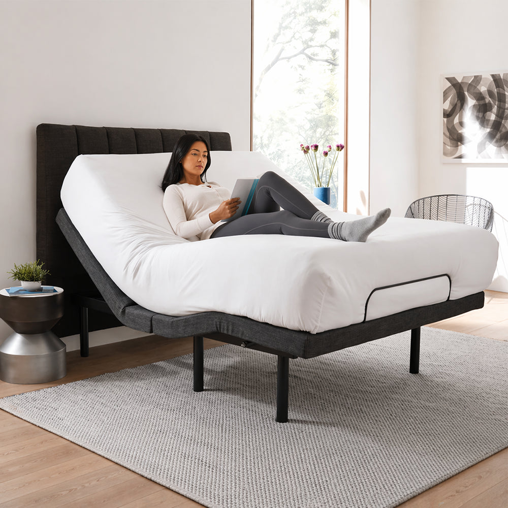 Woman sitting on an S1000 Adjustable Bed Base by SleepTone with the head section elevated for comfortable reading in a modern master bedroom.
