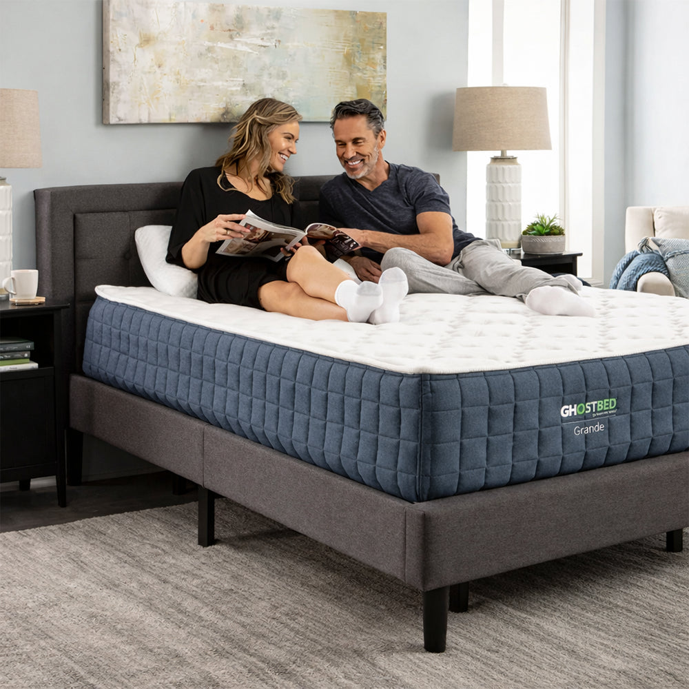 Couple sitting and reading on a supportive GhostBed Grande 14-inch medium mattress in a bright, contemporary bedroom.
