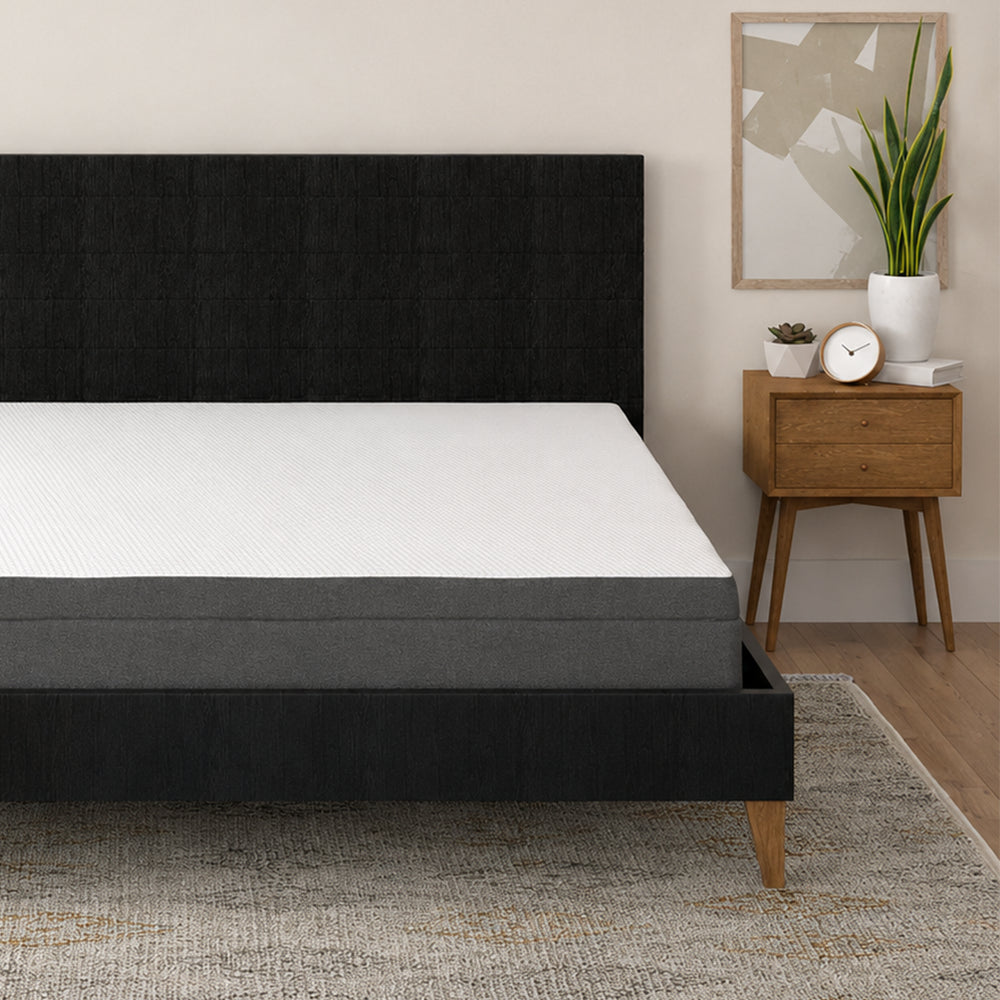 Perspective view of the South Bay Daisy firm memory foam mattress on a black upholstered platform bed beside a mid-century modern nightstand.
