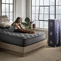 A woman stretching on a Corsicana Nights Bridge mattress in a loft bedroom, with the branded mattress-in-a-box shipping carton visible.
