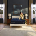 A woman sitting on a DreamCloud Ultra 15-inch Medium Hybrid Mattress in a modern master bedroom interior with designer lighting.