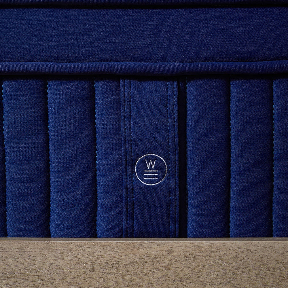 Macro detail of the circular Weekender brand emblem stitched into the vertical channel-quilted side of the medium plush hybrid bed.
